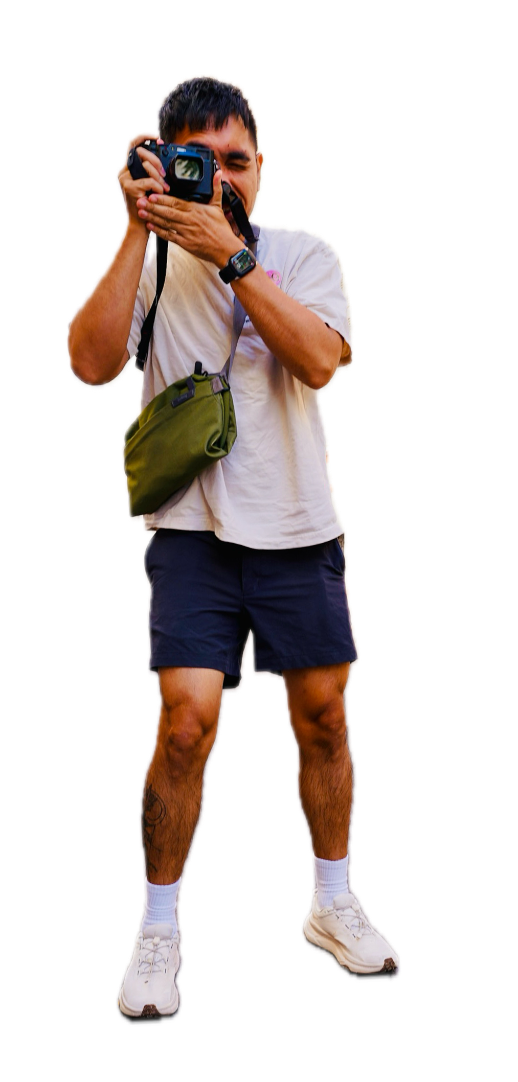 A man standing outdoors taking a photo with a camera, wearing a beige t-shirt, dark shorts, white sneakers, white socks, and a green bag across his chest.