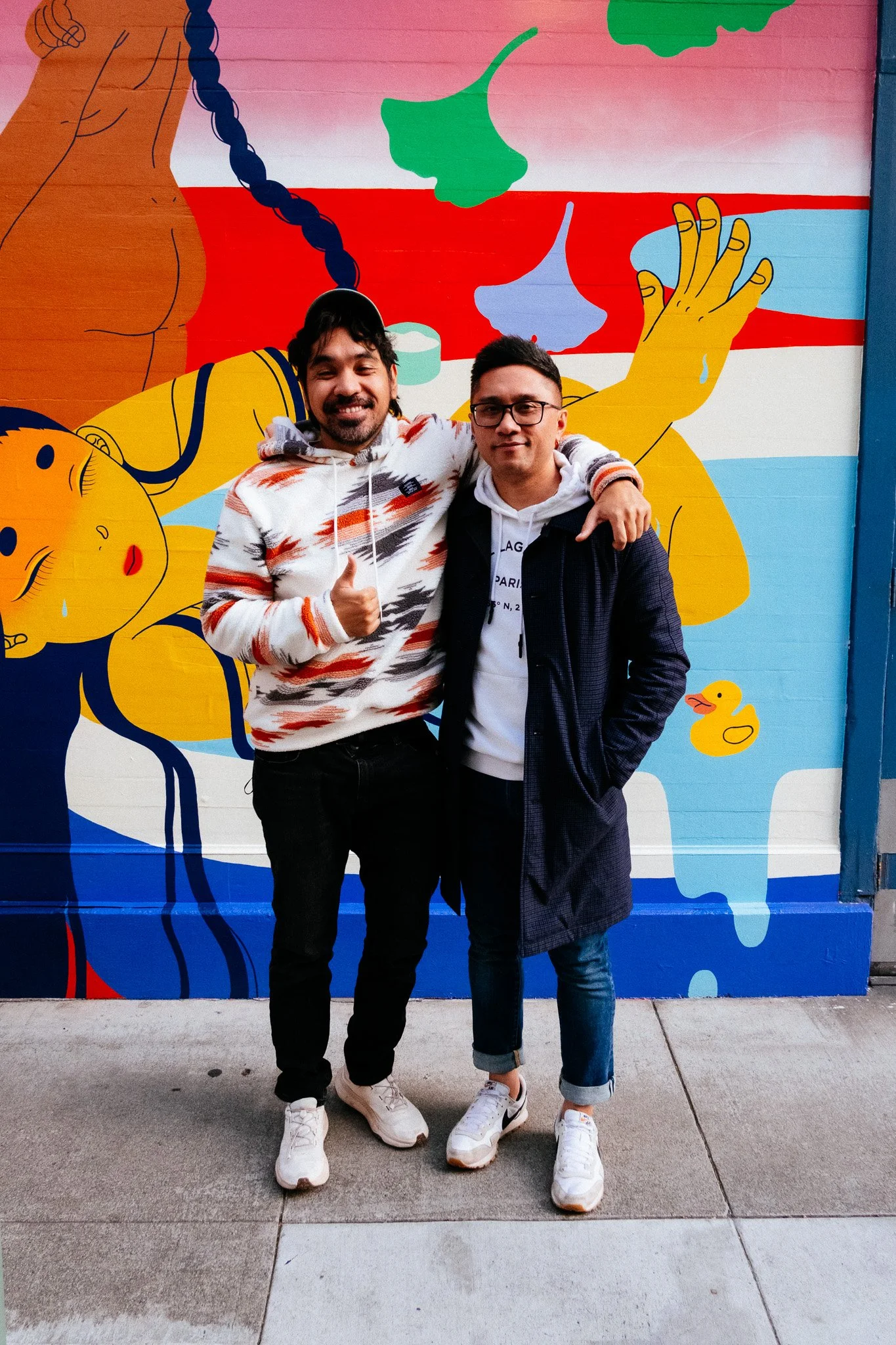 Two designers posing arm in arm in front of a colorful bathtub mural on a sidewalk