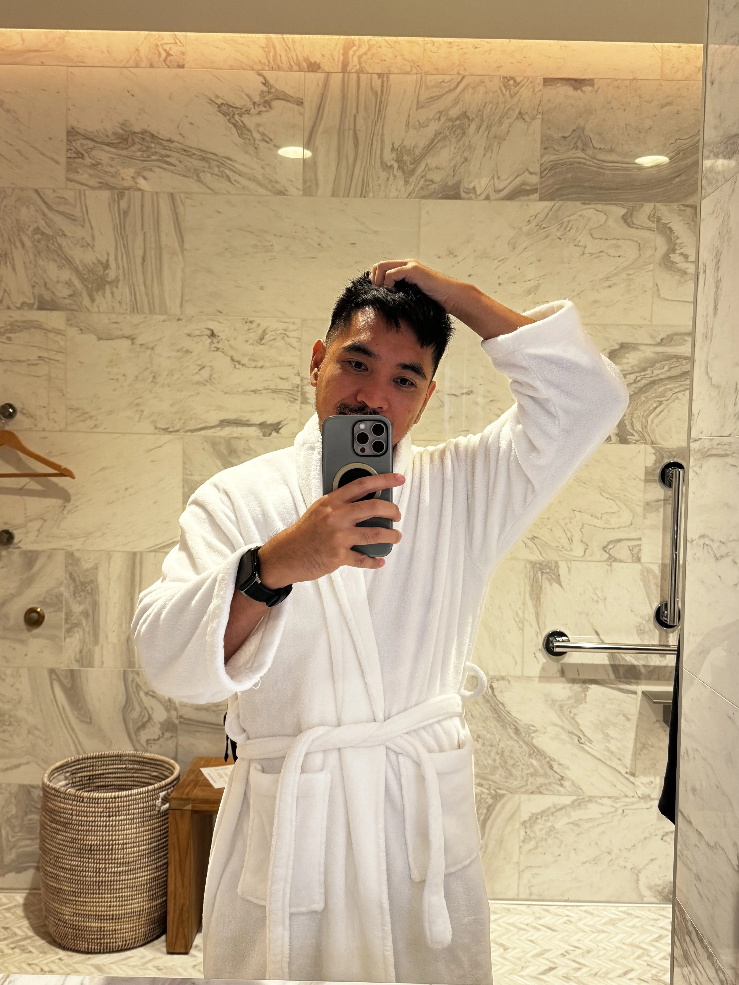 Mark taking a mirror selfie in a white bathrobe in a marble hotel bathroom