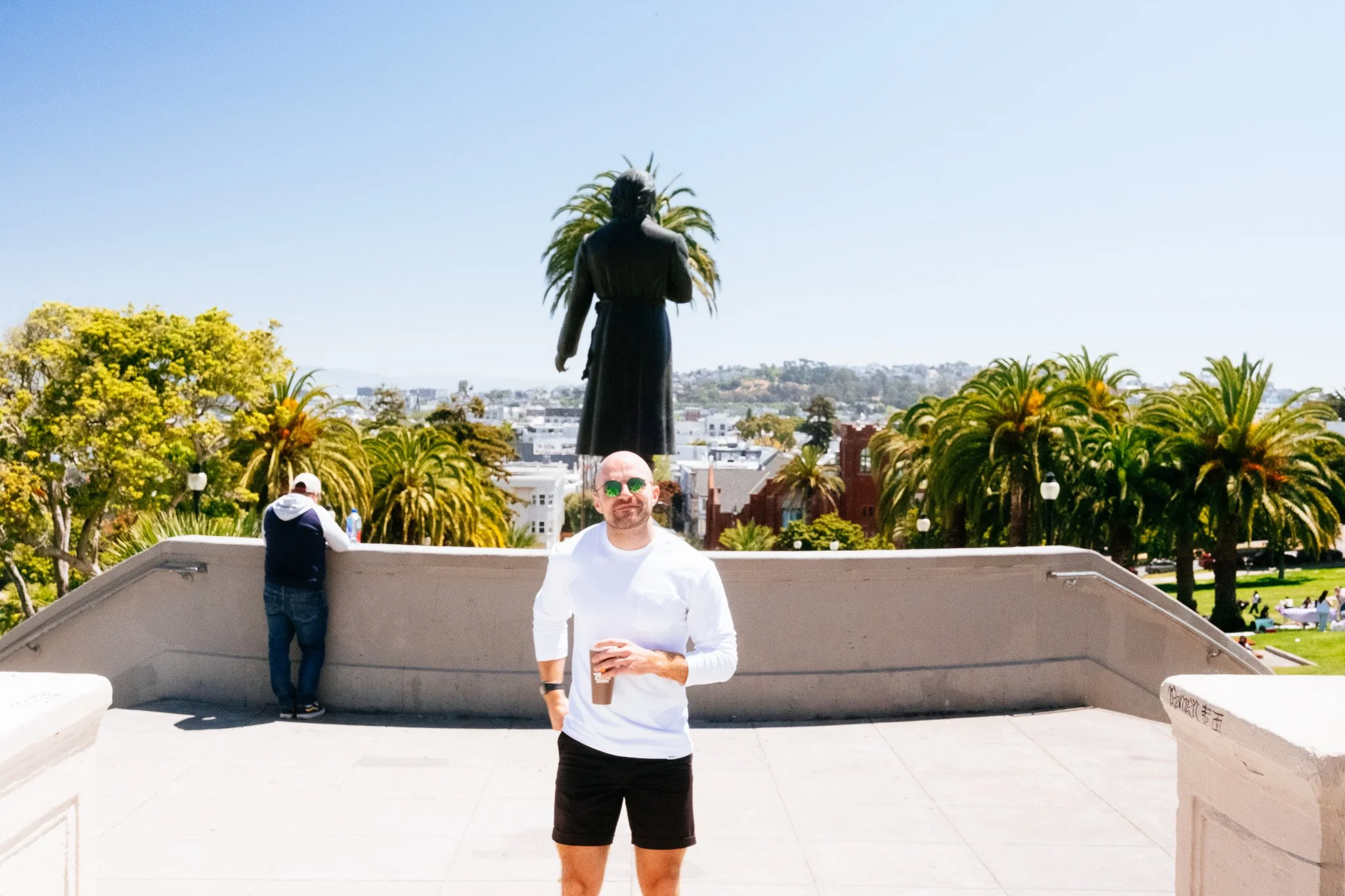 A friend holding an iced coffee on the overlook at Mission Dolores Park, statue and palms behind