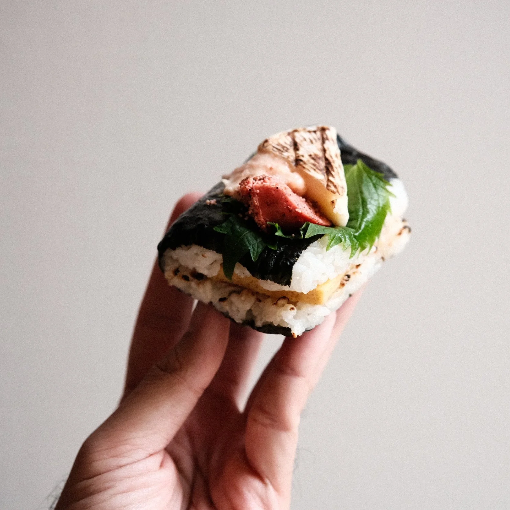 Hand holding an onigirazu rice sandwich filled with tamago, shiso and torched fish