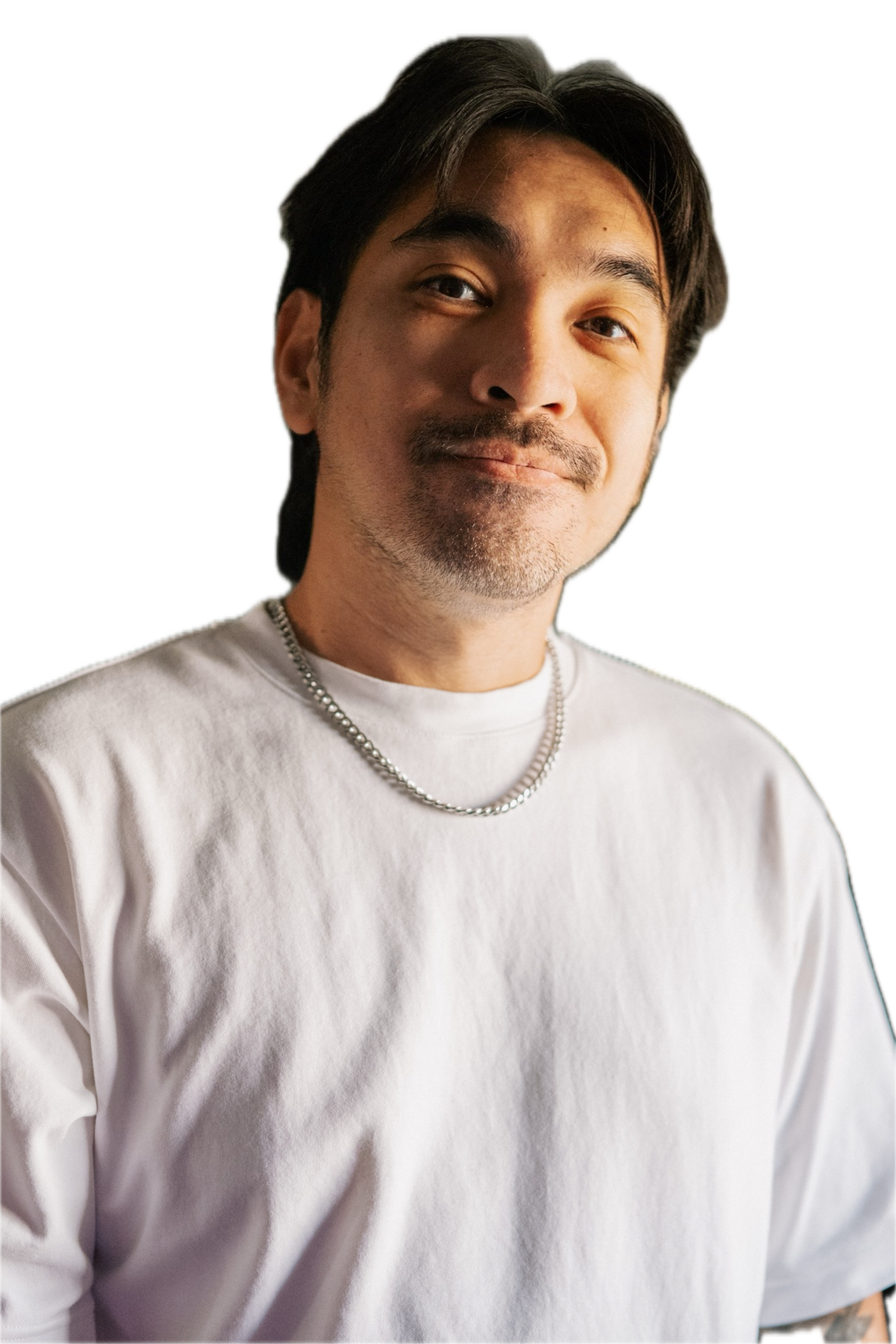 A young man with dark hair and a slight beard, smiling and looking at the camera, wearing a white t-shirt and a silver chain necklace.