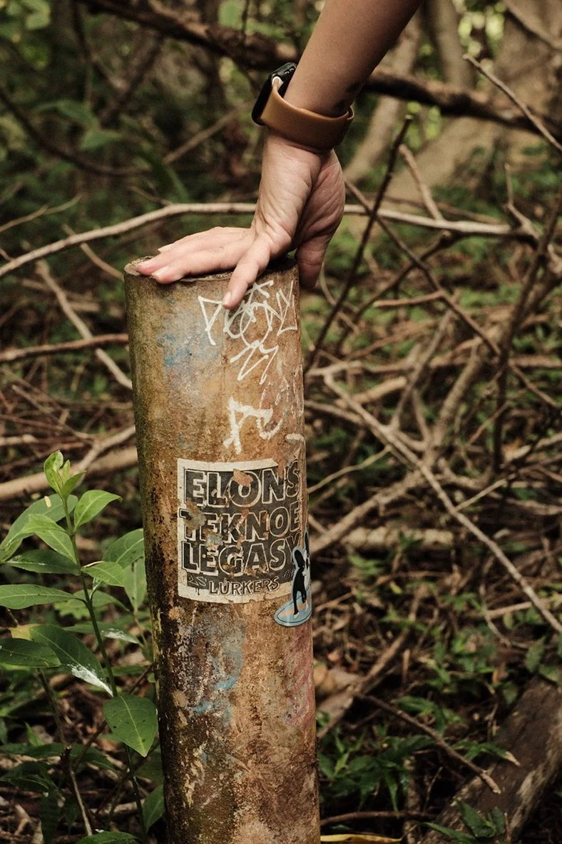 Hand resting on a sticker-covered concrete trail post in dense Oahu brush