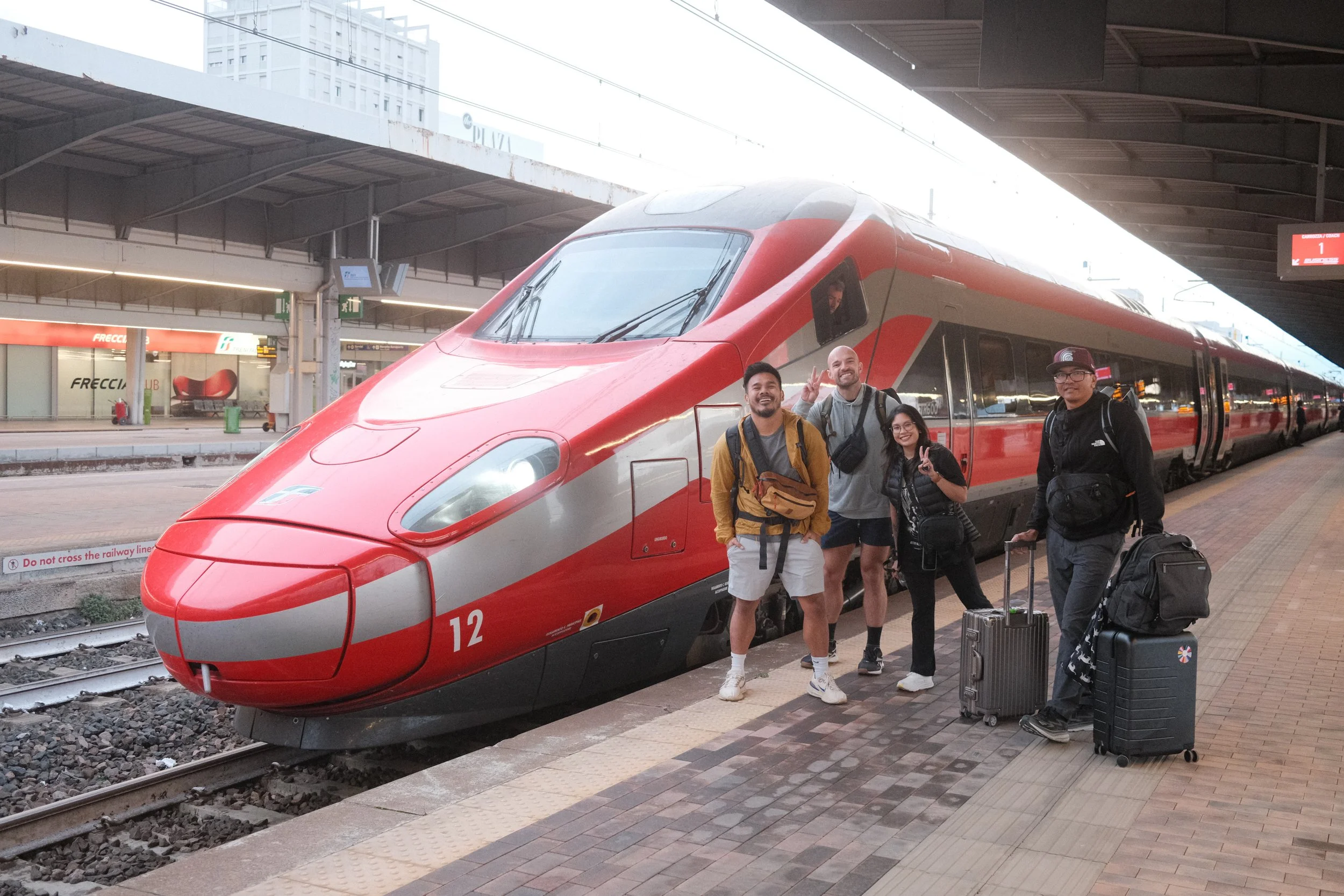 Four friends with luggage beside a red Frecciarossa high-speed train in Italy