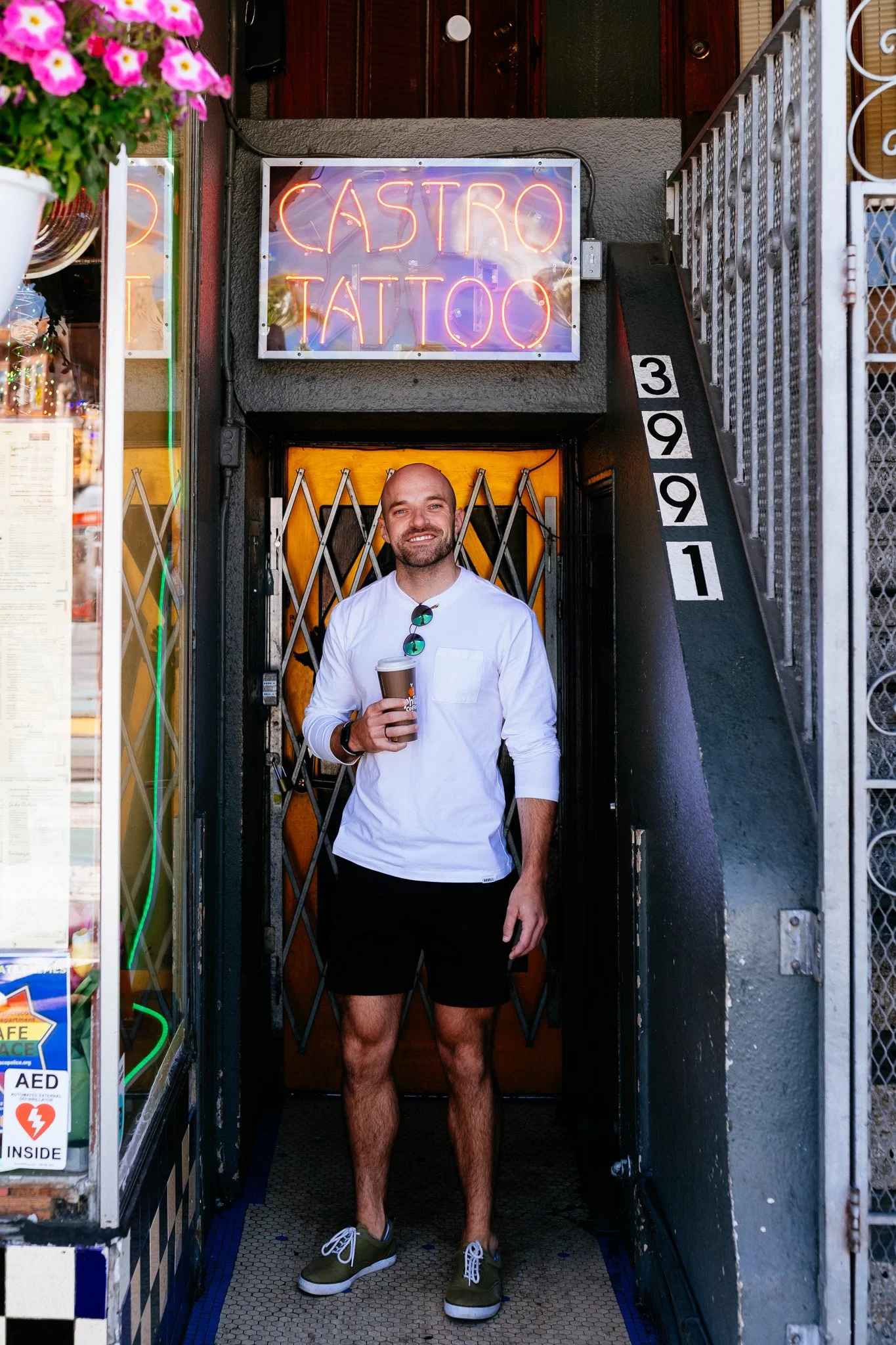 Smiling man holding coffee in a doorway beneath a Castro Tattoo neon sign